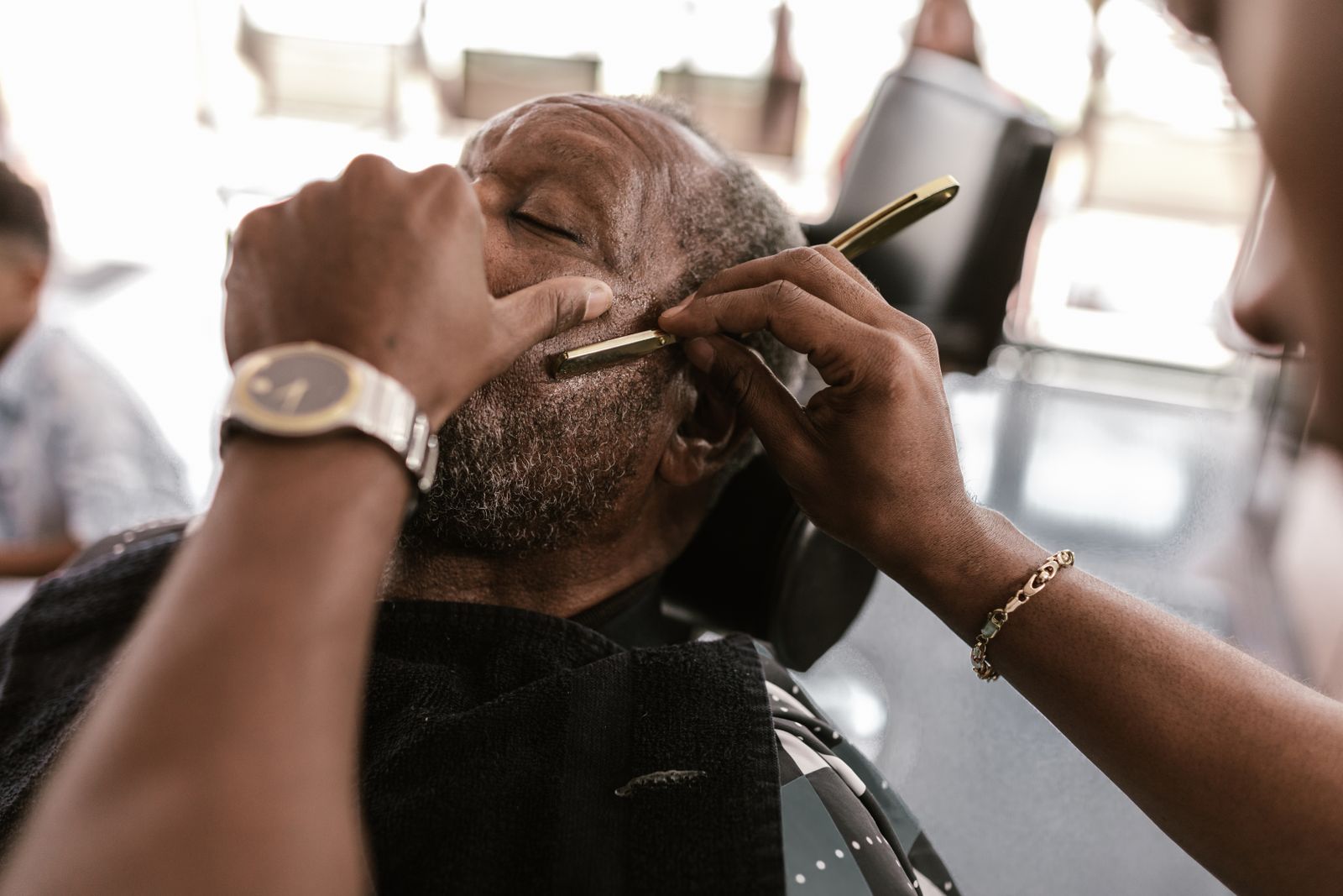 A barber shaping a seated customer’s beard with a razor.