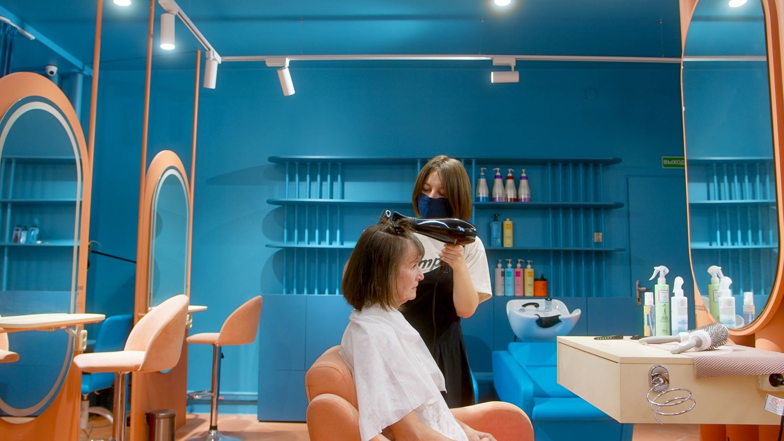 A hairstylist blow-drying a seated client’s hair in a salon.